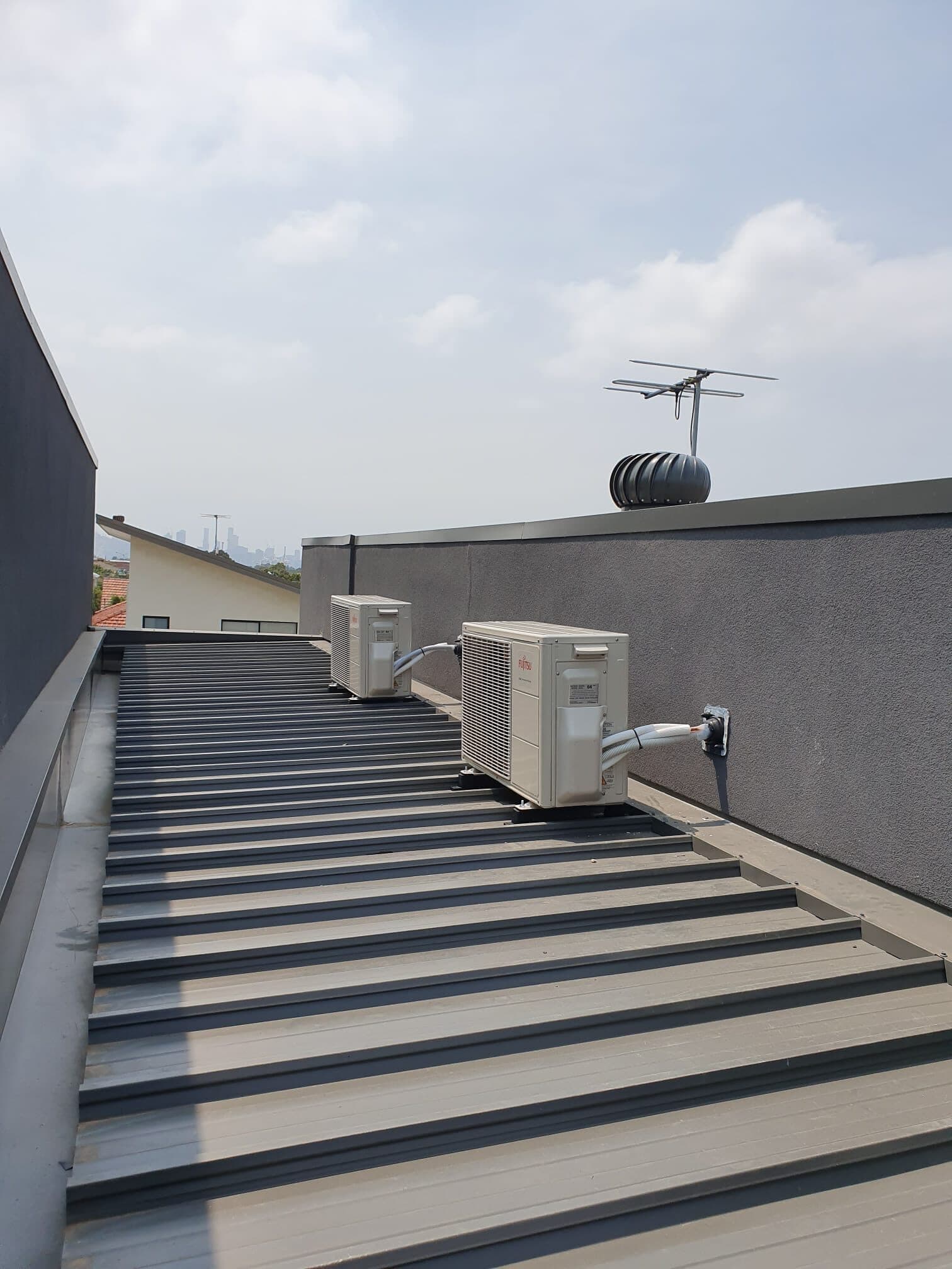 Two outdoor condensers on a flat rooftop in Abbotsford with neat conduit runs and the Melbourne city skyline behind