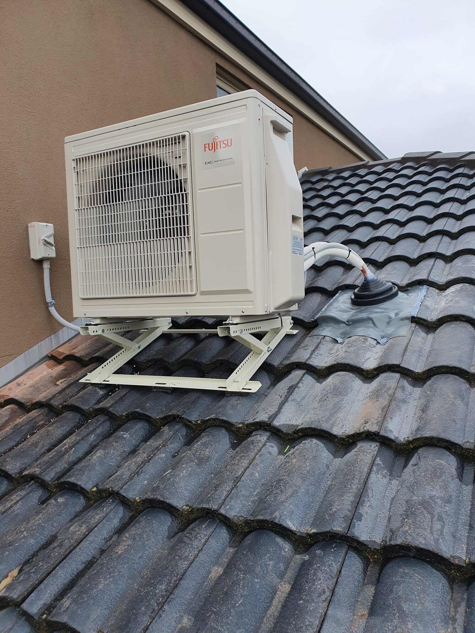 An outdoor condenser on a galvanised bracket fixed above a tiled house roof in Ascot Vale, with a tidy conduit run and flashed roof penetration