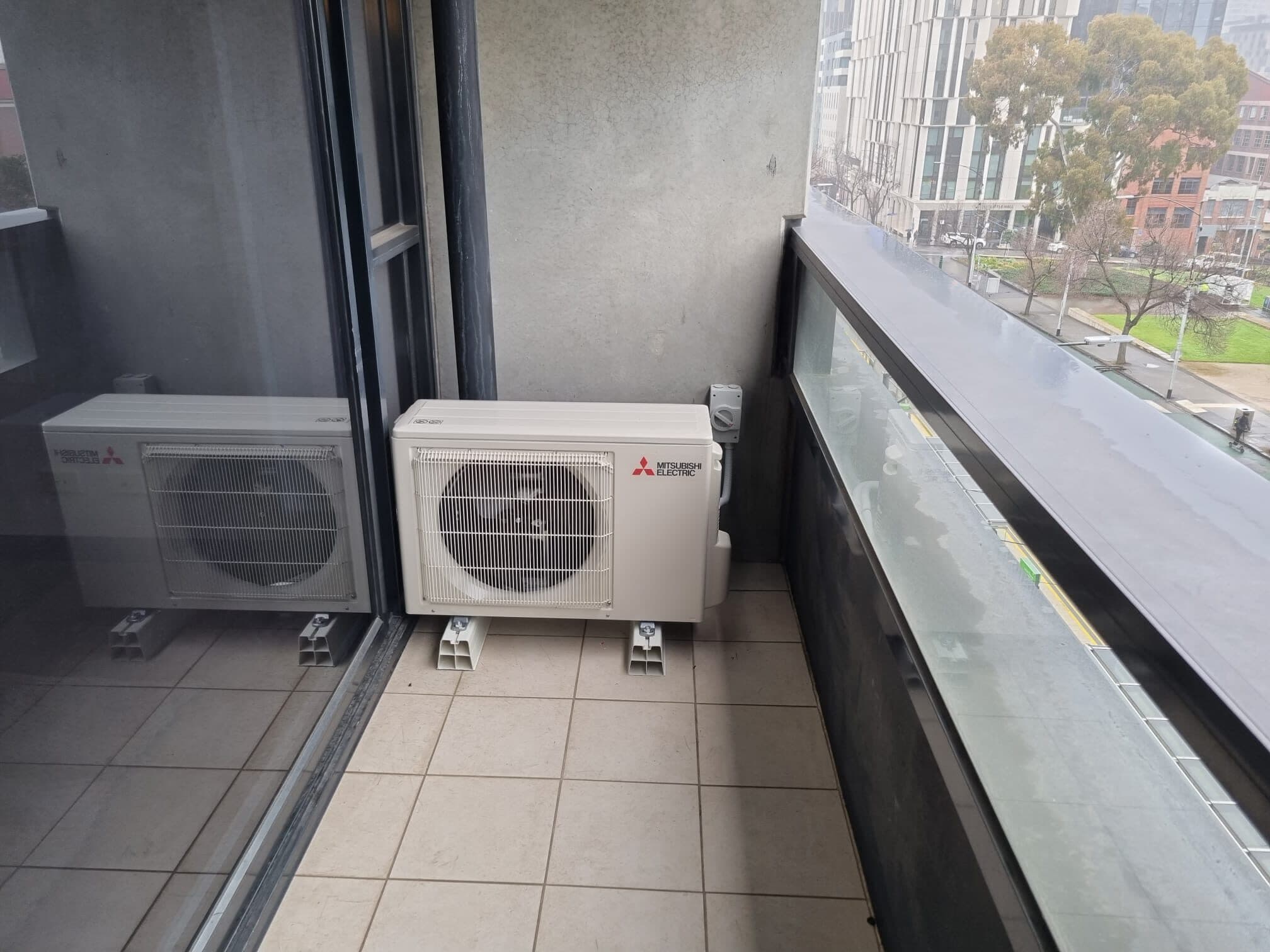 Two outdoor condensers set neatly on a tiled apartment balcony in Carlton with city towers and street trees behind