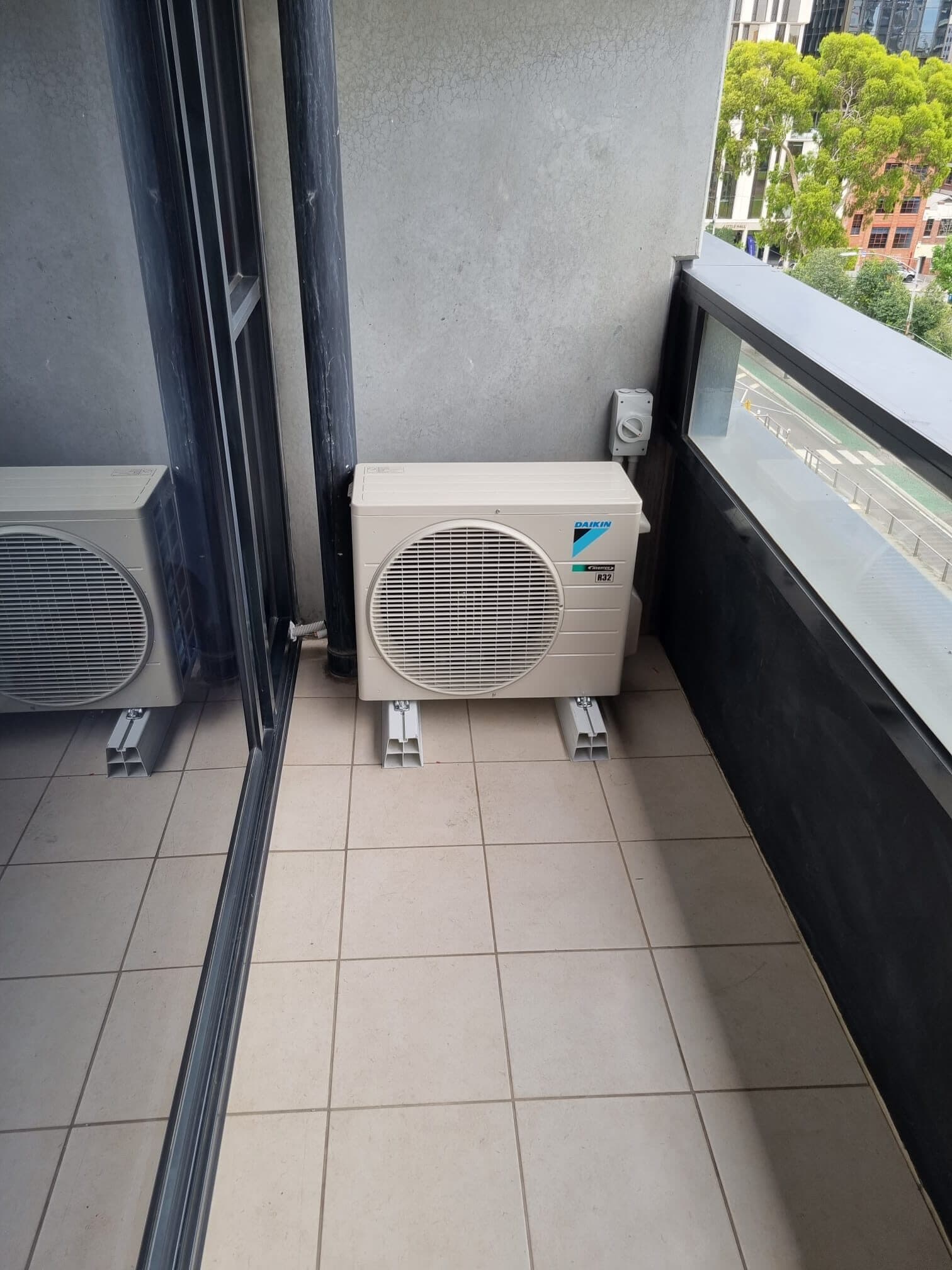 Two outdoor condensers on a tiled apartment balcony in East Melbourne with the street and rooftops visible through a glass balustrade