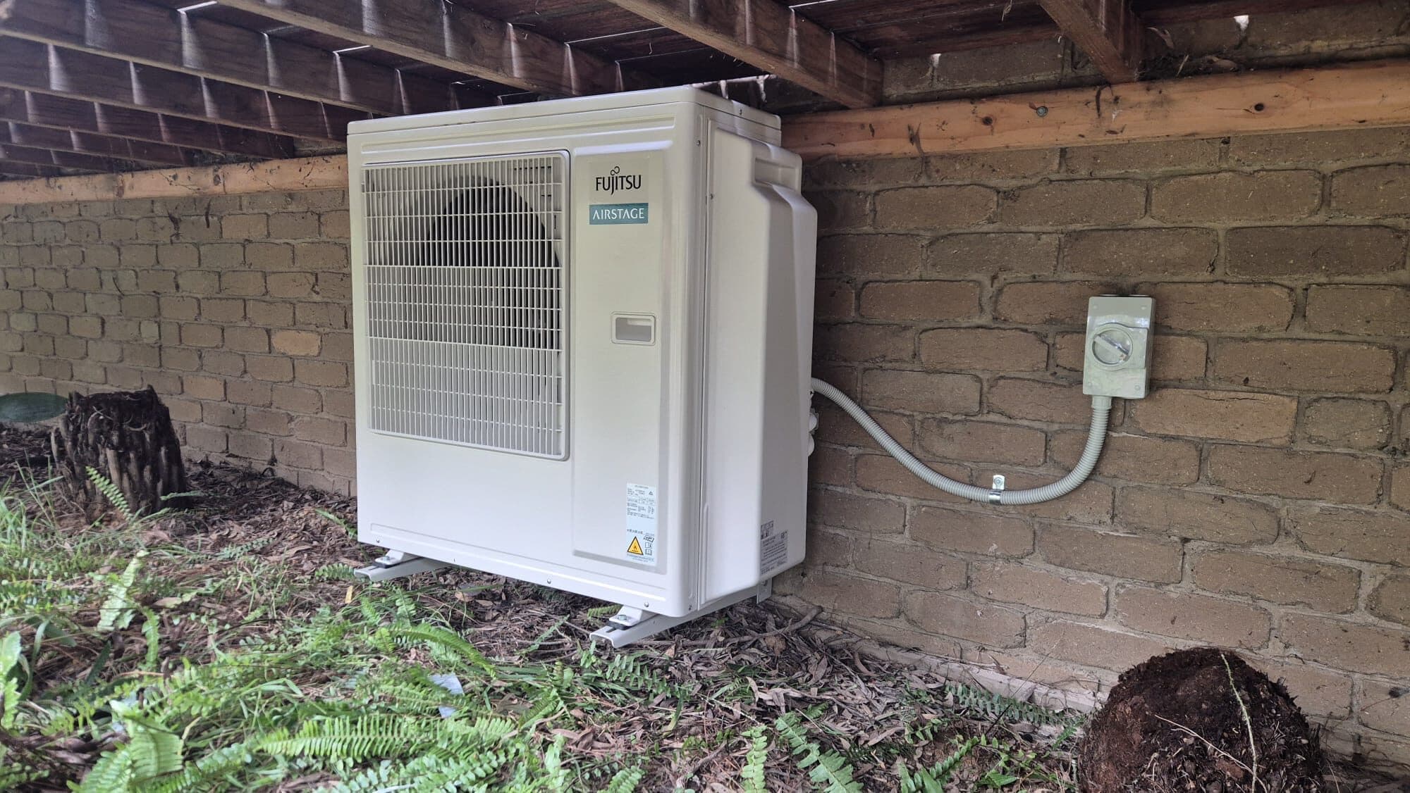 An outdoor condenser bracket-mounted on a painted brick subfloor wall in Eltham with a neat conduit run to an isolator