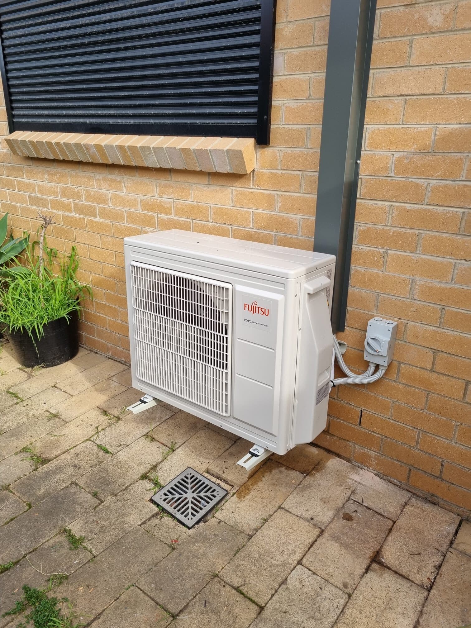 An outdoor condenser neatly bracket-mounted on a cream brick house wall beside a window in Essendon, with tidy conduit to the isolator