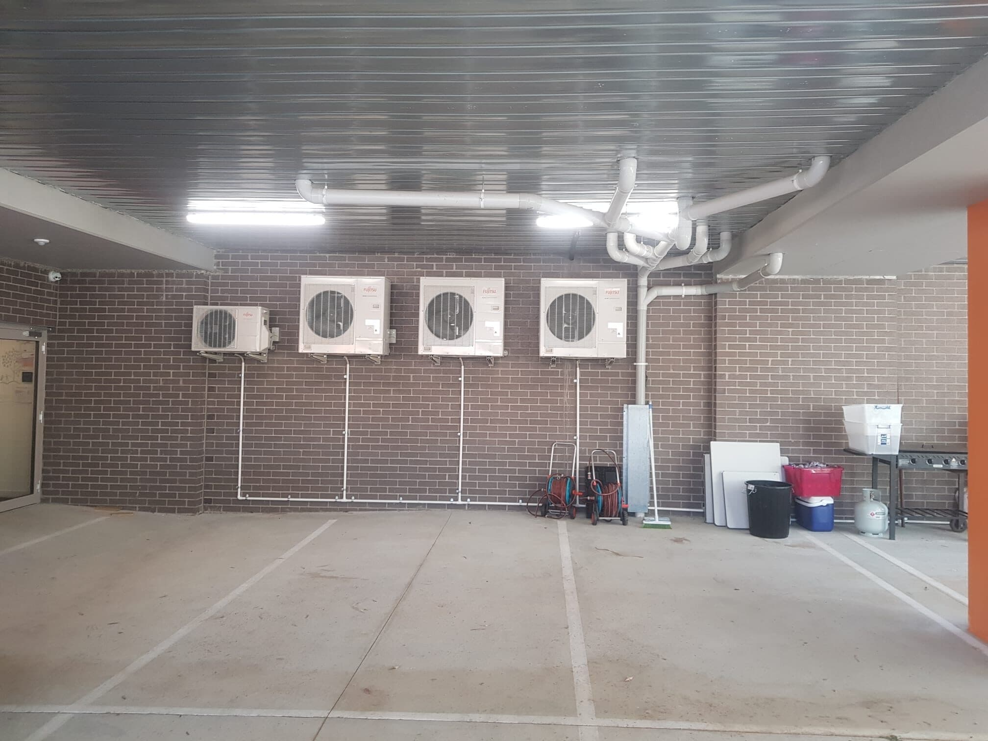 A row of four outdoor condensers wall-mounted under the undercroft of a childcare centre in Fairfield with overhead pipework to a manifold