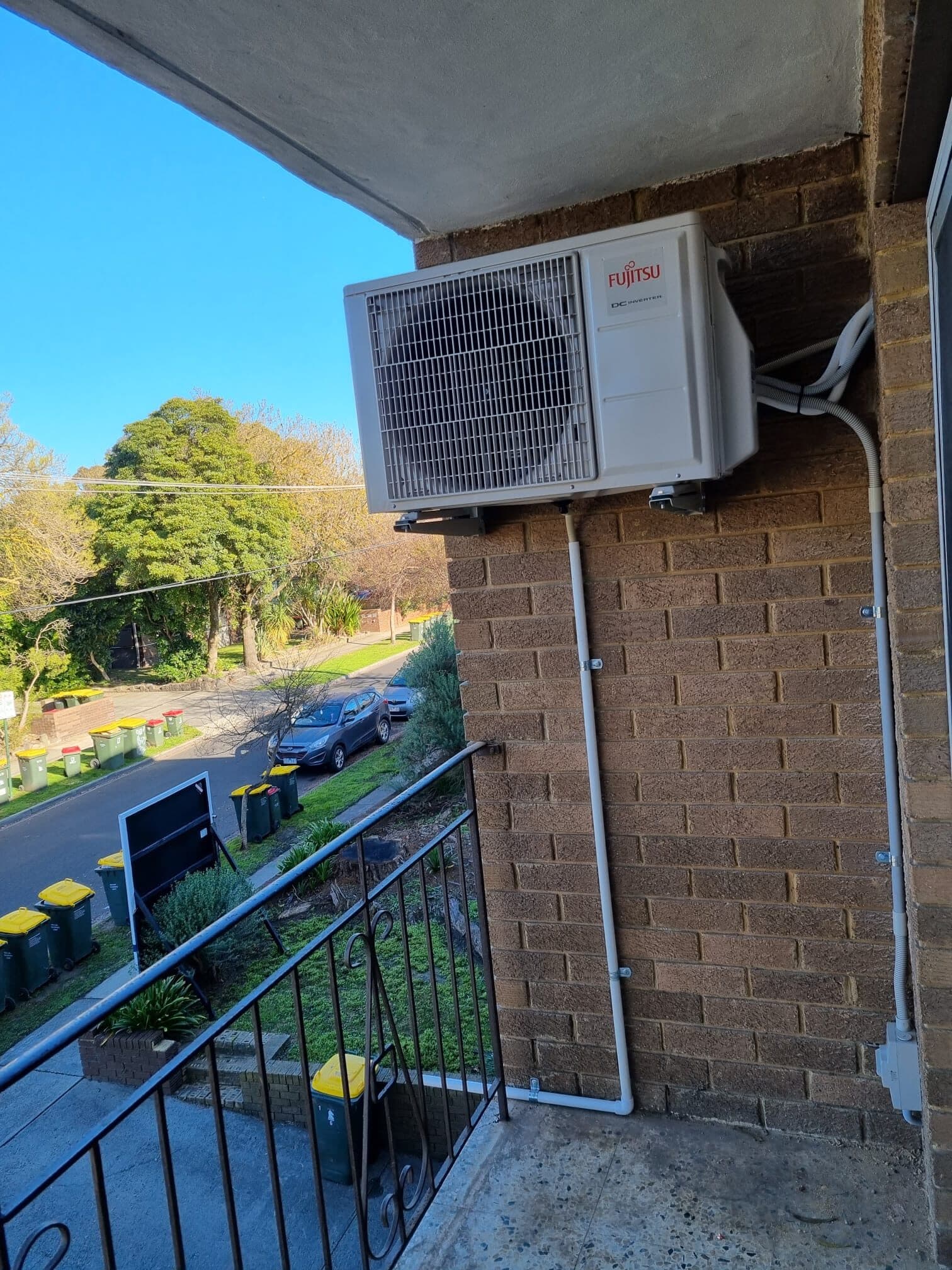 An outdoor condenser bracket-mounted on a brick balcony wall in Fitzroy with a tidy white conduit run and blue sky behind
