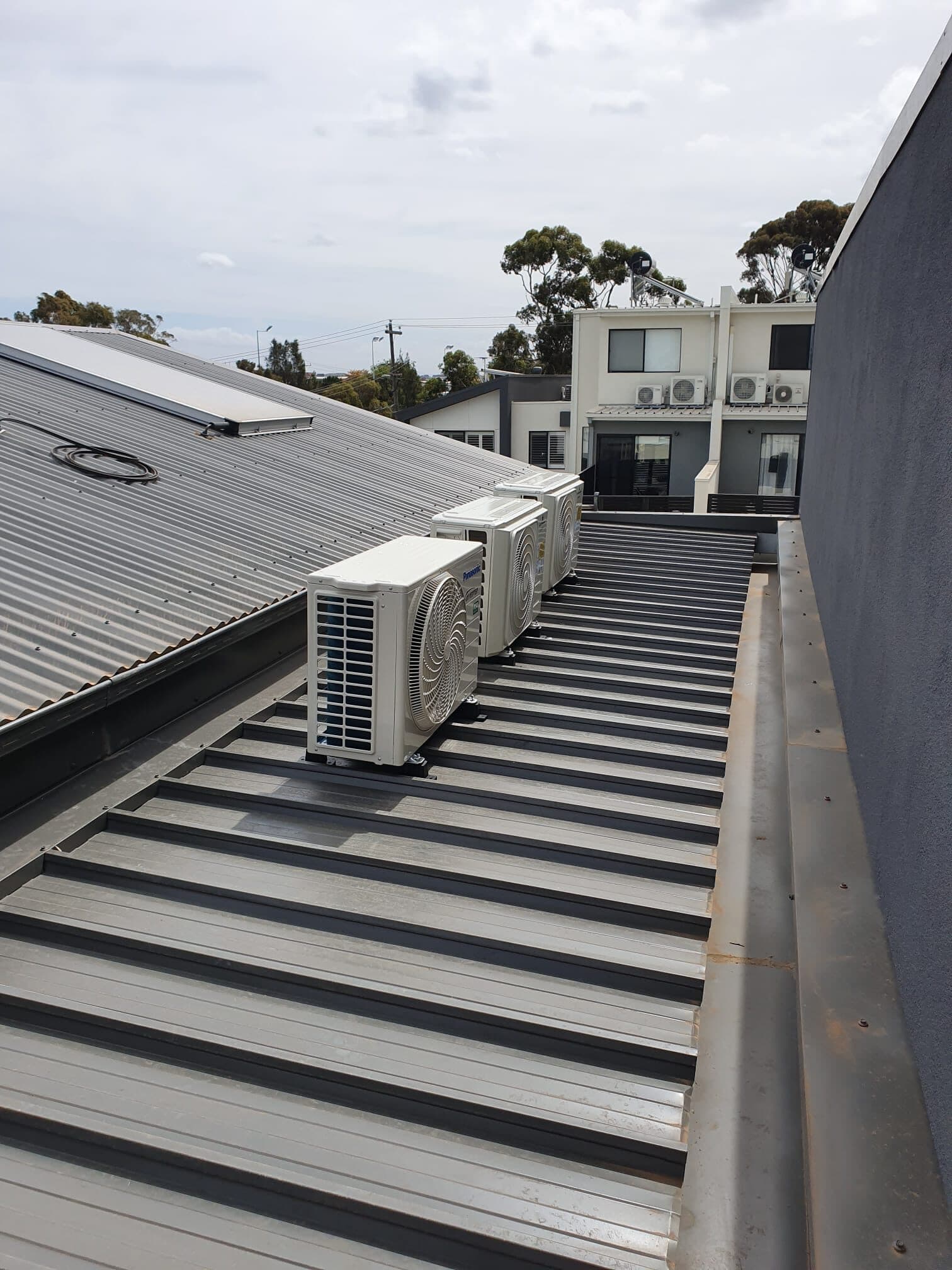 Three outdoor condensers lined up along the metal roof of a multi-dwelling townhouse development in Glen Waverley, with a second block behind