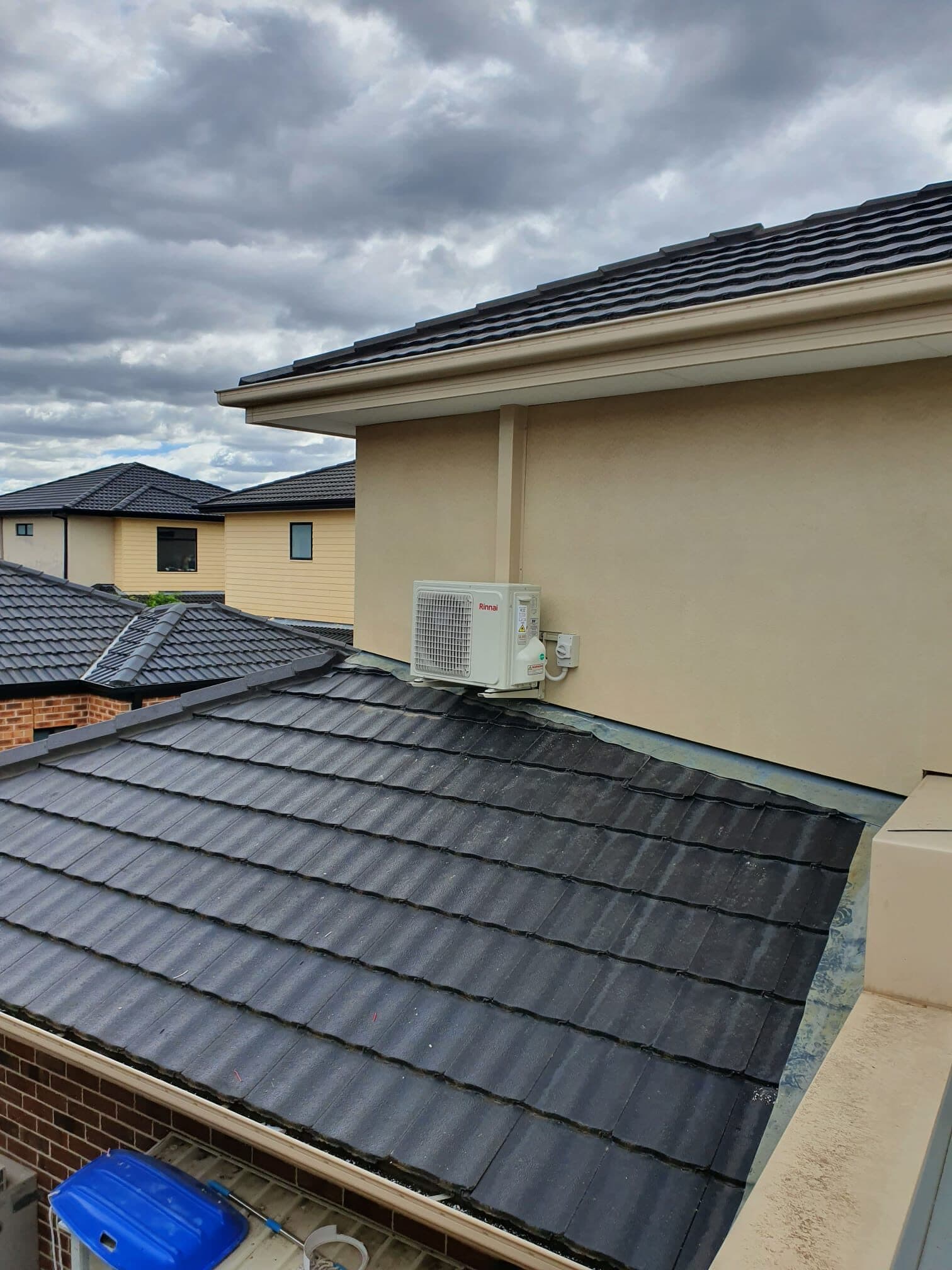 An outdoor condenser mounted on the rendered upper wall of a new townhouse in Glenroy, with rows of matching new rooftops behind
