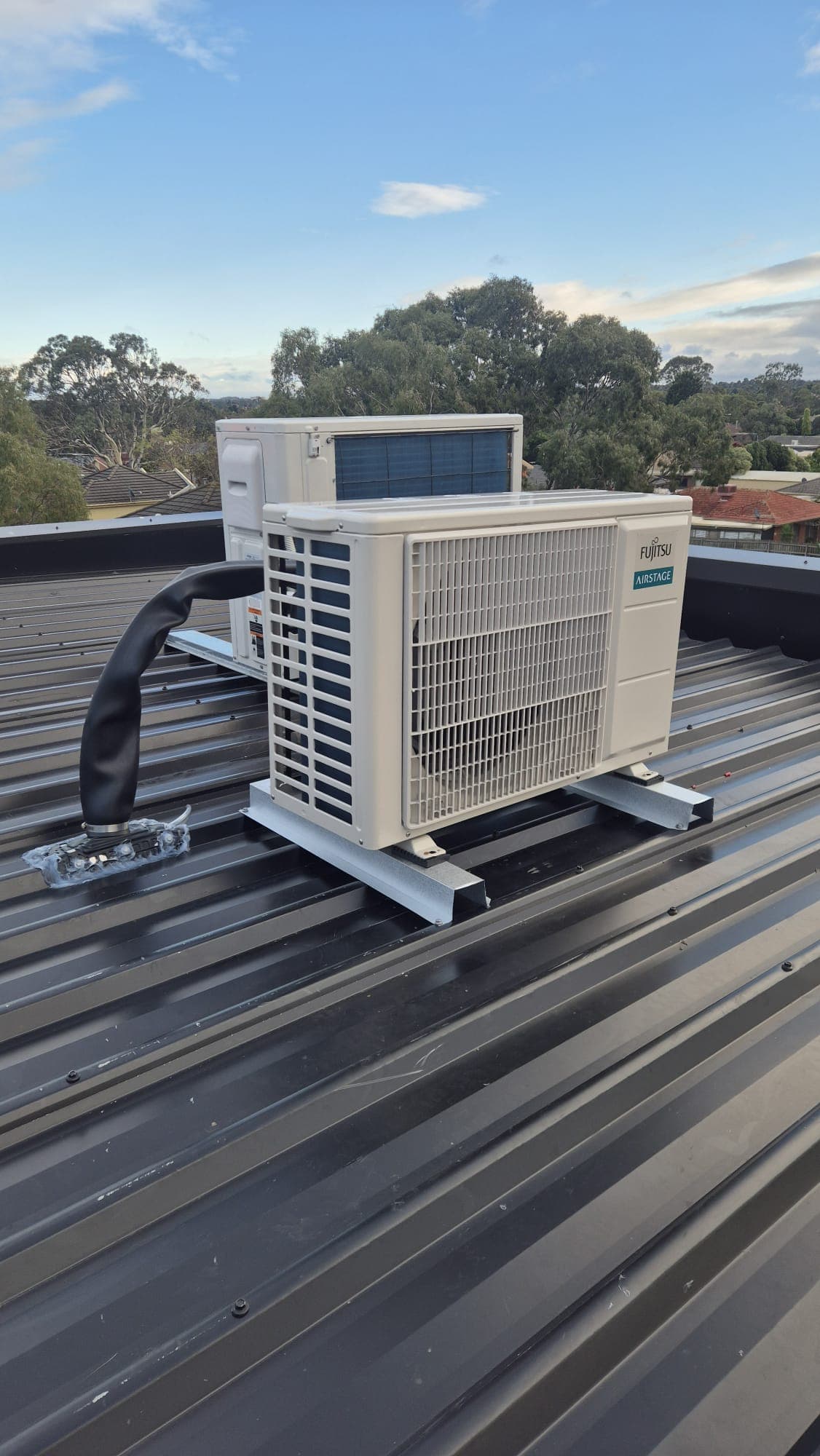 An outdoor condenser on a metal roof in Mill Park with black elephant-trunk pipe protection against a clear blue sky