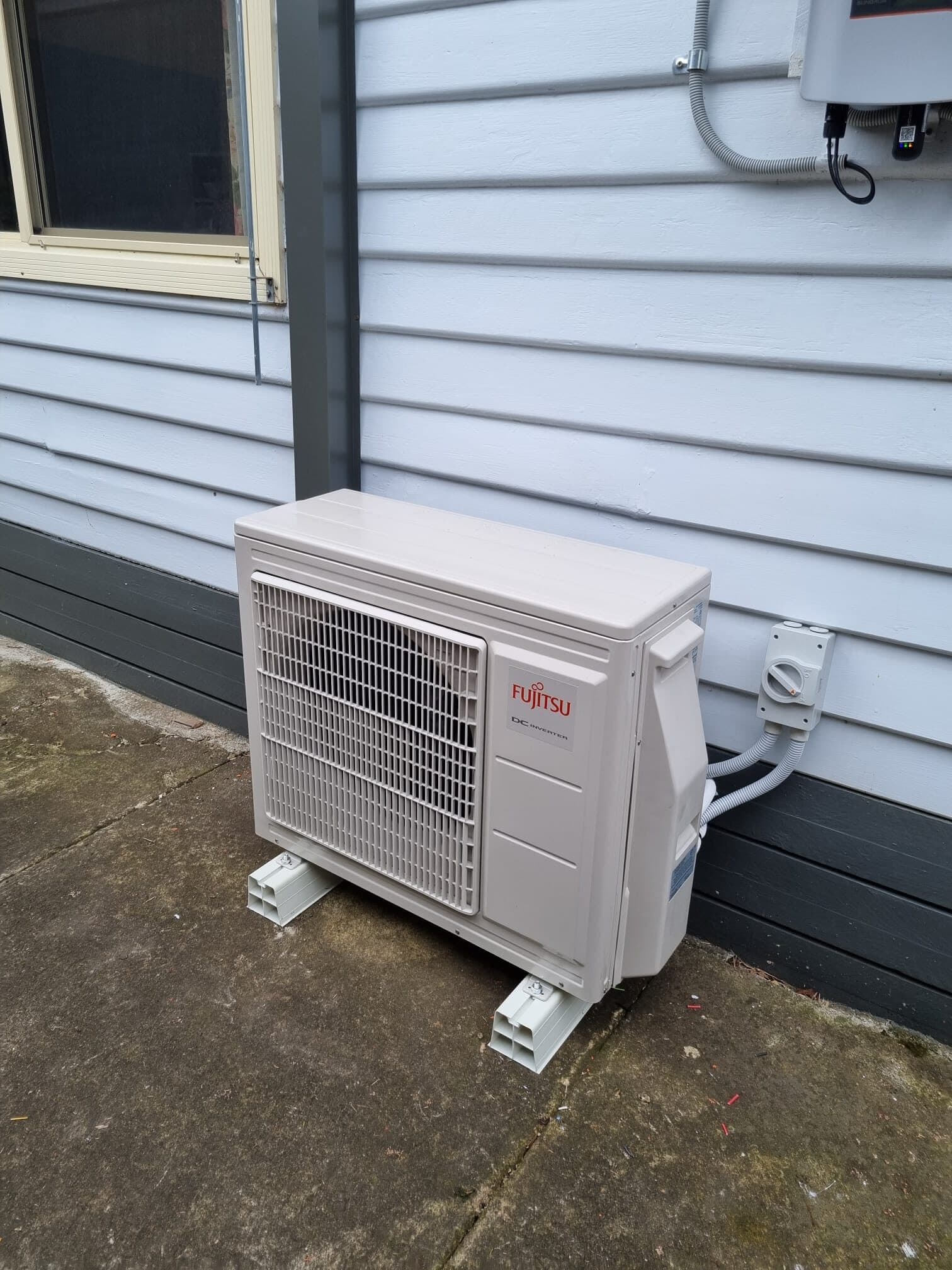 An outdoor condenser ground-mounted on alloy feet against grey weatherboard in Richmond, isolator switch above