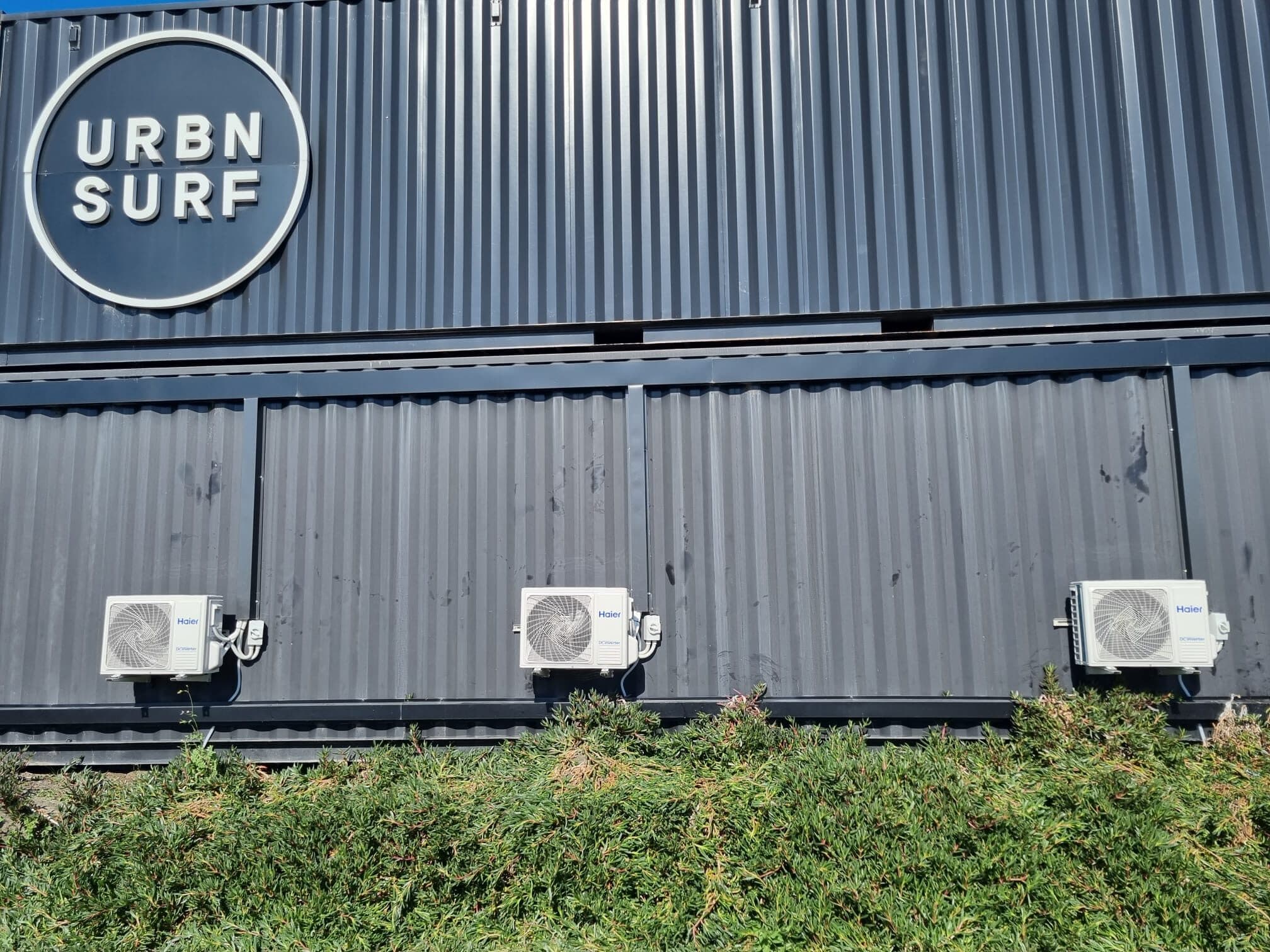 Three outdoor condensers neatly mounted on the dark corrugated facade of Urbn Surf in Tullamarine, the Urbn Surf sign above