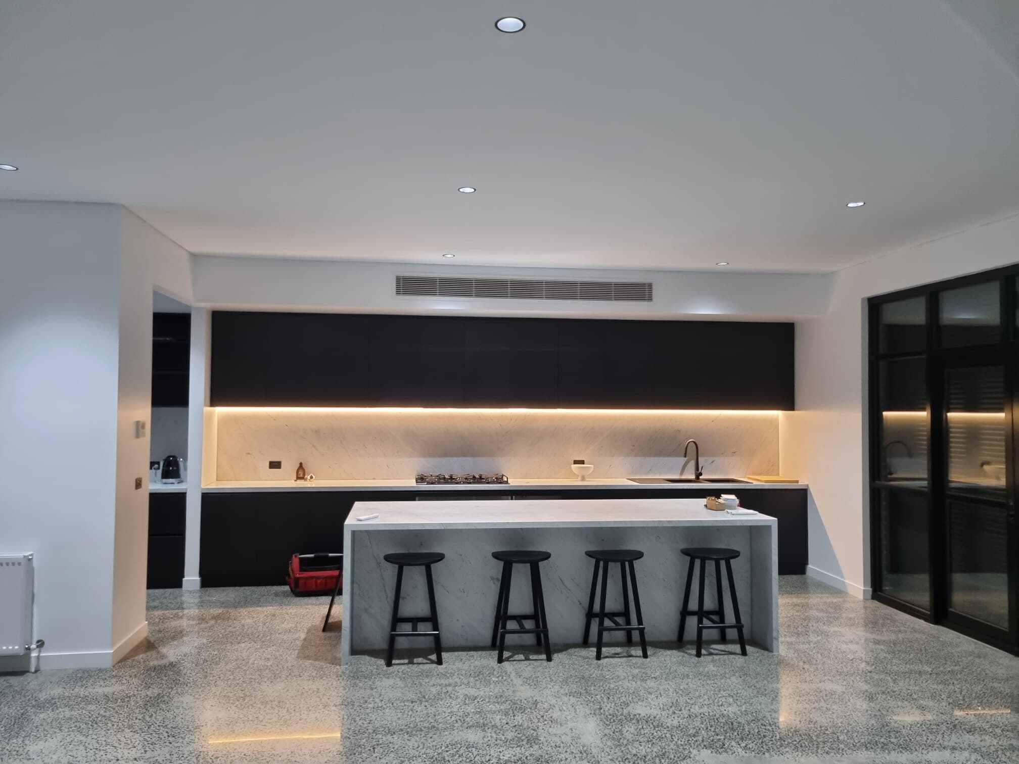 A ducted bulkhead grille set discreetly into the soffit above the cabinetry of a high-end kitchen in Wheelers Hill, with a polished concrete floor and marble island