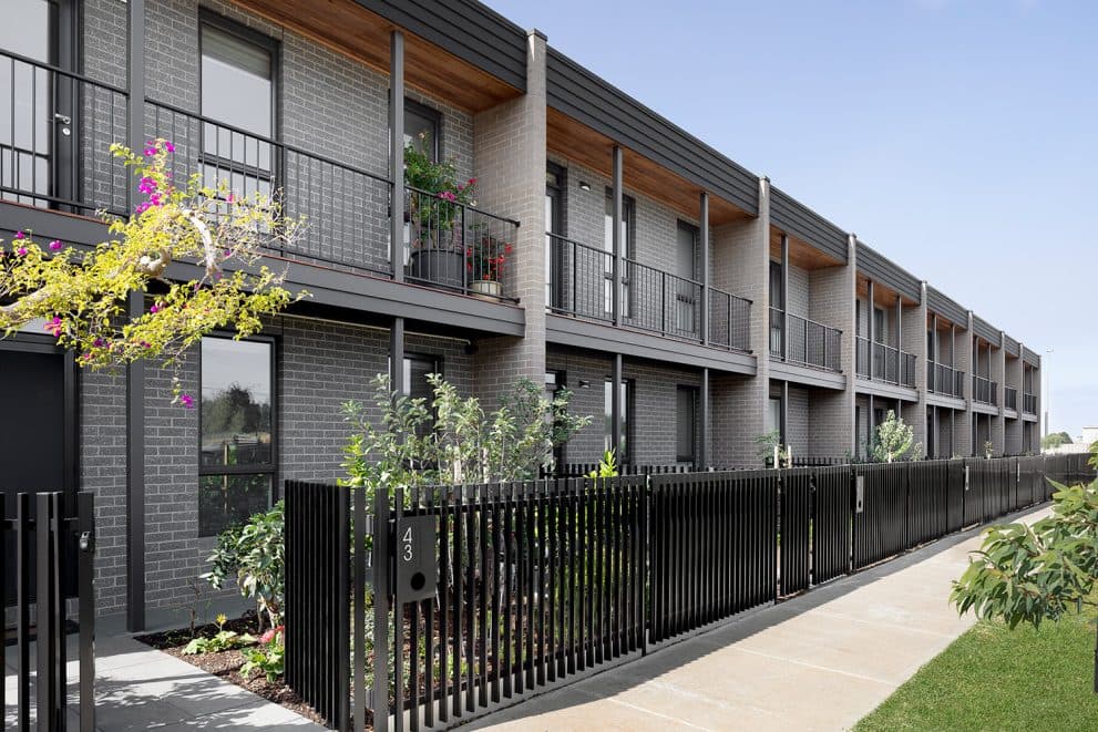 Street view along the Luma townhomes, charcoal brick facades with timber-lined balconies behind a black batten fence